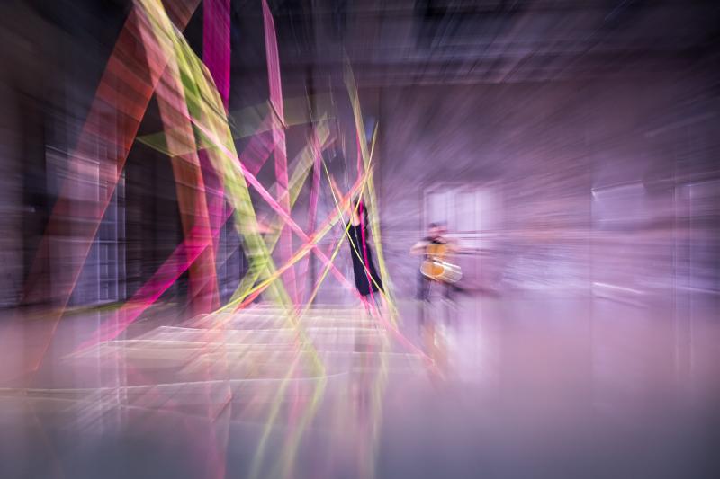 immagine usata per la comunicazione dell'evento: una foto sfuocata on fasci luminosi colorati di una stanza con all'interno sullo sfondo due persone una suona un contrabbasso e la donna danza
